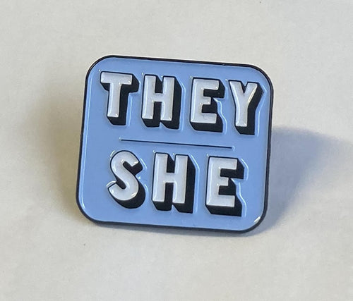 A blue enamel pin with the words 'They' and 'She' featured in capital letters. This pin is square with rounded edges.